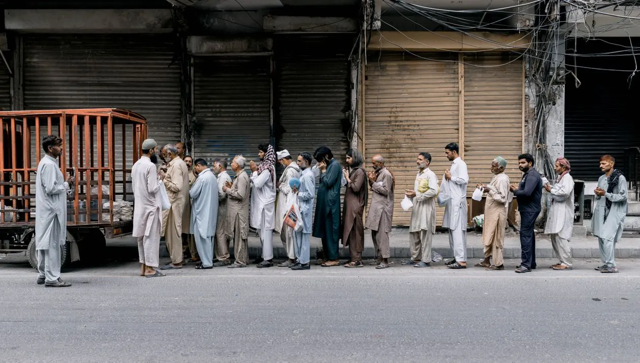 Men standing in a long queue outside closed shops in Faisalabad while waiting to receive relief goods.