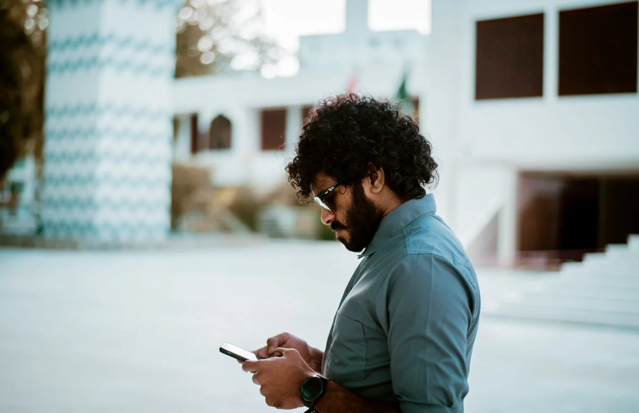 A young man with curly hair checking his smartphone outdoors in a bright courtyard.