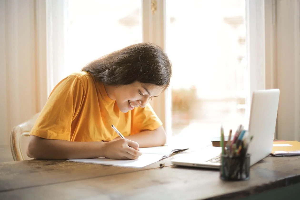 A student writing study notes at a desk beside her laptop in bright daylight